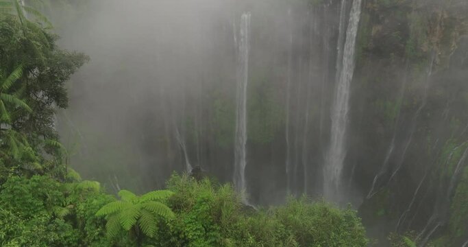 Aerial Tilt Up Shot Of Waterfall Flowing On Rock Formations Against Cloudy Sky - Java, Indonesia