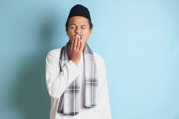 Portrait of attractive Asian muslim man in white shirt yawning with hand covering his mouth. Isolated image on blue background