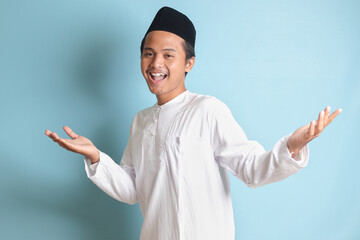 Portrait of Asian muslim man in white koko shirt with skullcap pulling hands towards camera,...