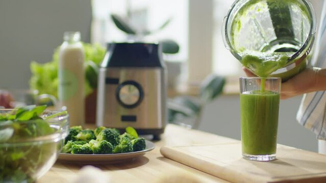 Cropped selective focus shot of hands of woman filling glass with freshly made green smoothie from blender in kitchen at home