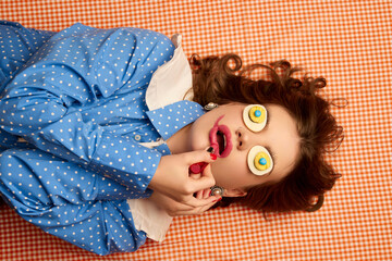 Doing makeup. Weird people concept. Young girl lying on table with eggs on her eyes over peach color background. Vintage, retro style. Food pop art photography.