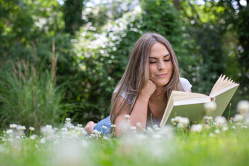 young blond woman lies in the garden between many daisies, reads a book and smiles