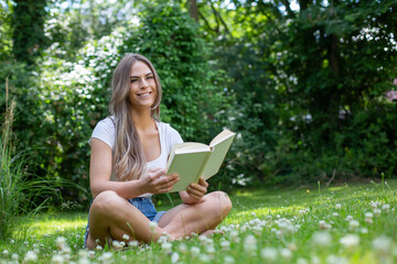 Fototapeta premium Young blond woman sits cross-legged in the garden between many daisies, reads a book and smiles