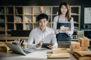 Young Asian SMEs woman and man looking to camera, standing among several boxes in modern  office.