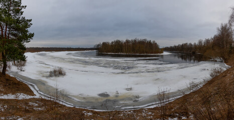 Winter. Overcast and cloudy. A sharp bend in the river, ice, gullies in the ice. Islet with a birch grove. Panoramic photography