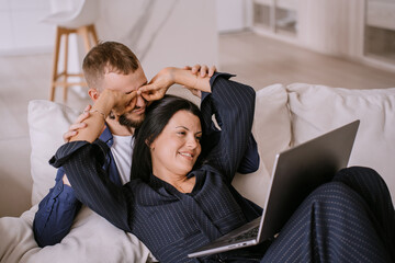 Young hispanic couple having fun home laying on couch with laptop talks with friends via internet. Brunette woman makes video call, doing glasses for boyfriend by hands. Weekend, holiday, valentine's.