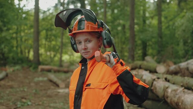 Portrait of a female logger stands in the forest, a young specialist woman in protective gear holds an axe in her hands and works on deforestation.