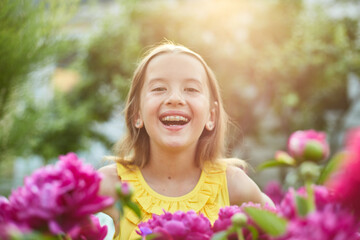 Happy little girl with braces in the garden in bushes of peonies - obrazy, fototapety, plakaty