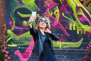 A woman wearing leather clothes takes a selfie against a wall with graffiti