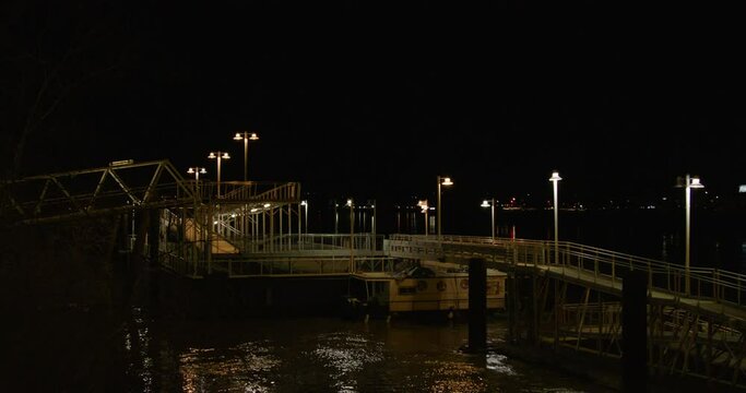 A lot of lampposts help to light this bridge that connects a dock in the darkness. The current of the river flows past the docked houseboat. It is a wide shot.Traffic passes by in the background.