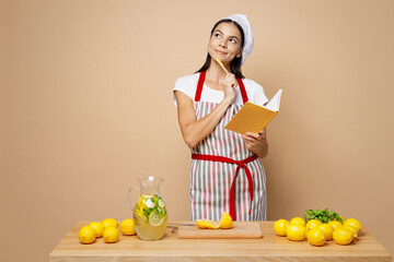 Obraz premium Young minded housewife housekeeper chef latin woman wears apron toque hat work at table kitchenware cutting lemons make lemonade writing in cook book isolated on plain pastel light beige background.