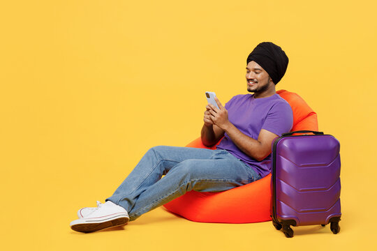 Traveler Sikh Indian man wear turban dastar purple t-shirt sit in bag chair use mobile cell phone at suitcase isolated on plain yellow background. Tourist travel in free time. Air flight trip concept.
