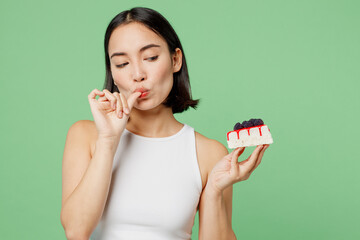 Young happy fun woman wear white clothes holding in hand pice of cake dessert lick fingers isolated on plain pastel light green background. Proper nutrition healthy fast food unhealthy choice concept.