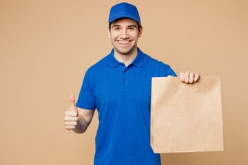 Delivery guy employee man wear blue cap t-shirt uniform workwear work as dealer courier hold brown clear blank craft paper takeaway bag mock up show thumb up isolated on plain light beige background.