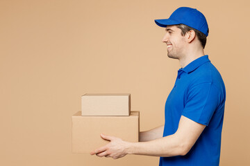 Side profile view fun delivery guy employee man wear blue cap t-shirt uniform workwear work as dealer courier hold stack cardboard blank boxes isolated on plain light beige background Service concept - obrazy, fototapety, plakaty