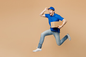 Fototapeta na wymiar Full body side view delivery guy employee man wear blue cap t-shirt uniform workwear work as dealer courier jump high hold cardboard box run isolated on plain light beige background. Service concept.
