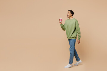 Full body young man of African American ethnicity wear green sweatshirt hold takeaway delivery craft paper brown cup coffee to go isolated on plain pastel beige background. People lifestyle concept.