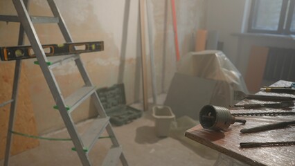 Room under renovation. Ladder with water level, wooden table with metal nozzles for perforator or electric drill. Spiral borers for drilling and bowl for cutting holes for socket.