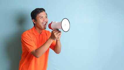 Portrait of attractive Asian man in orange shirt speaking louder using megaphone, promoting...