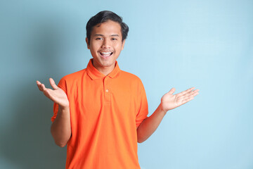 Portrait of attractive Asian man in orange shirt spreading his hands sideways and holding two things, demonstrate products. Welcoming someone. Isolated image on blue background