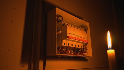 High voltage automatic breaker switch, close up in the dark. Candle illuminates a white electrical panel with many wires, automatic switches, breakers, residual current devices, fuses. Blackout.