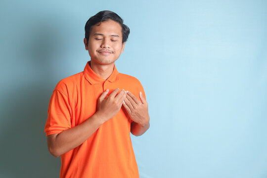 Portrait of calming Asian man in orange shirt placing hand on chest and feeling peaceful. Mental health day concept, expresses sympathy and love, smiles positively. Isolated image on blue background