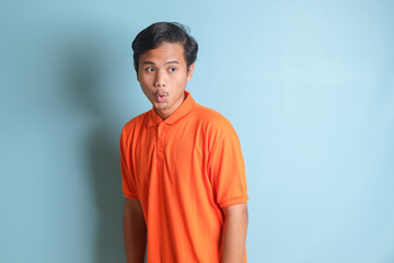 Obraz premium Portrait of surprised Asian man in orange shirt standing against blue background, showing shocked expression and looking aside