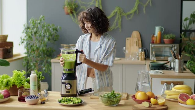 Medium long shot of young pretty woman pouring green smoothie from blender into glass in kitchen at home