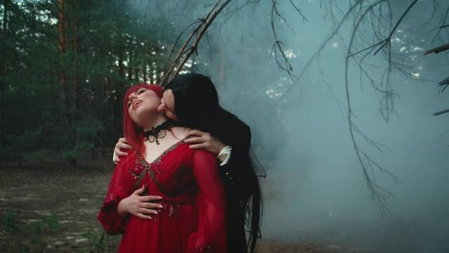 Goth man vampire in black tailcoat hugs bites kisses neck drinks blood of woman. art bloody drops dripping down from skin red lips. Happy lady fantasy couple two evil face. Red dress fog forest tree