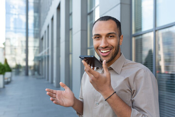 Outdoor portrait of young smiling African American man outside office building, man recording audio message smiling and looking at camera, businessman satisfied with work of AI voice assistant.