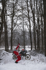 Happy motorcyclist in a Santa Claus suit shows likes. Winter forest with falling snow. Touring motorcycle in the background. The concept of New Year's holidays. Vertical photo.