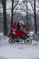 Naklejka premium A motorcyclist in a Santa Claus suit funny jumping. Winter forest with falling snow. Motorcycle in the background. The concept of New Year's holidays and sales. Vertical photo.