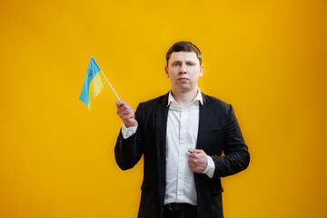 Focused sad businessman wearing suit holding Ukrainian flag standing over yellow studio background and looking camera