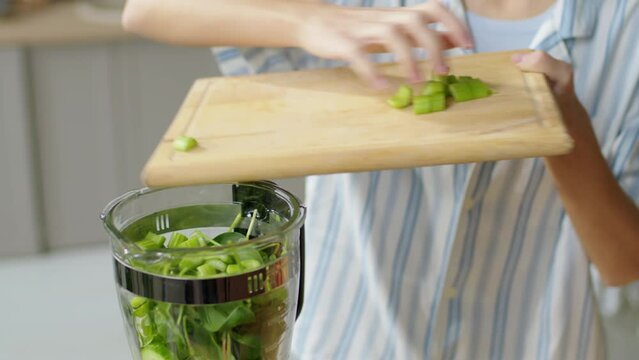 Cropped tilt up shot of woman putting fresh celery pieces into blender before making green smoothie at home