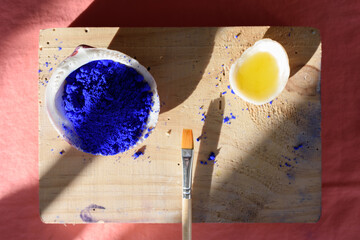 Pigmento azul cobalto en una concha, aceite en otra concha y pincel sobre madera, que a su vez está colocada sobre una tela cobriza, y todo ello recibiendo la proyección de luces y sombras.  - obrazy, fototapety, plakaty