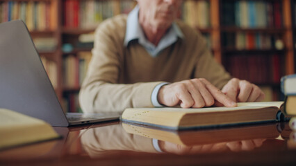 Senior historian reading a book and typing notes on laptop, working on an article