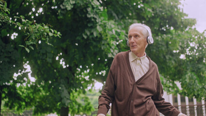 Naklejka na ściany i meble Cheerful senior man in headphones dancing during walk in park, good mood