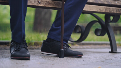 Feet of man sitting on bench in park and leaning on walking cane, close-up