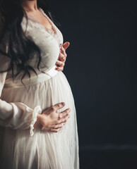 Beautiful pregnant woman with dark long hair in a white dress with lace hugs or ;her belly with her hands on a black background side view closeup