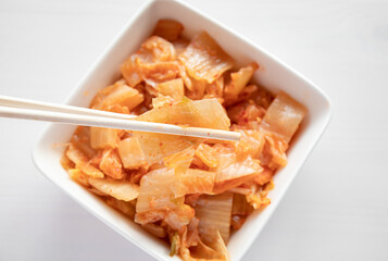 Selective focus on bamboo chopsticks with fermented kimchi cabbage salad and white food bowl on background. 