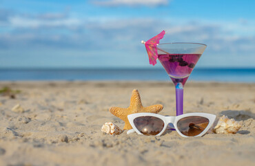 Glass of cocktail on sandy beach and sunglasses next to sea