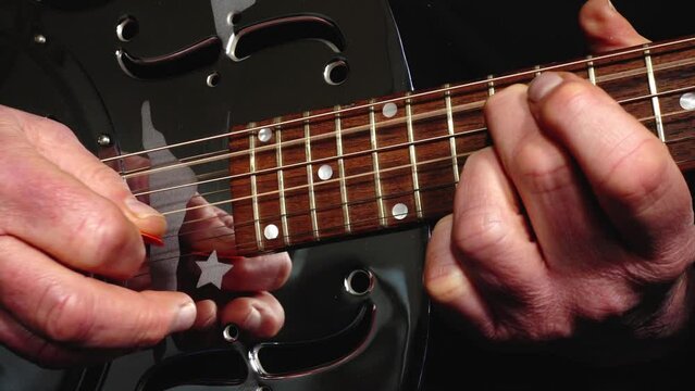 Closeup of a man&rsquo;s hands playing a shiny, metal body, resonator acoustic guitar, picking strings with one hand, while making chord shapes on the fretboard with the other.