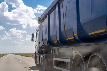 Large truck for bulk materials, gravel, grain and products driving along highway against blue sky on summer day. Modern heavy blue dumper truck moving on two lane suburban highway road, side back view © sommersby