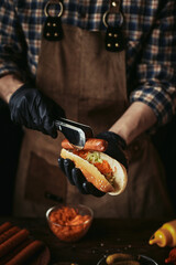 A guy in a leather apron makes a hot dog. The chef in black gloves puts a sausage in a bun.