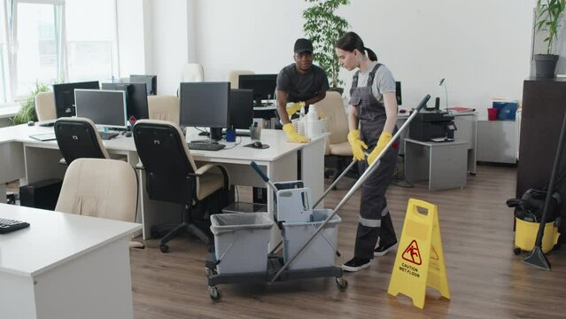 Slow motion long shot of young Black man and Caucasian woman wearing uniform cleaning modern open plan office getting rid of trash and dust