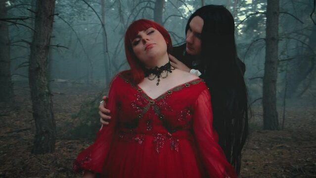 Goth man vampire in black tailcoat hugs bites kisses neck drinks blood of woman. art bloody drops dripping down from skin red lips. Happy lady fantasy couple two evil face. Red dress fog forest tree