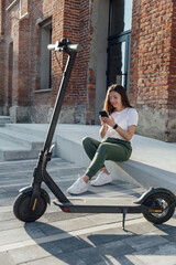 Young female sitting on urban stairs and speaking on smartphone near electric scooter