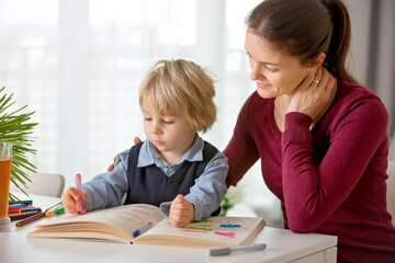 Fototapeta na wymiar Cute preschool child, blond boy, filling some homework in a work book and coloring, mother helping him
