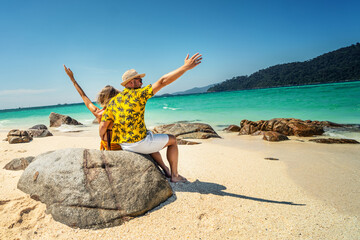 Happy Couple Enjoying Beautiful seascape, sitting with hands up on the rock, on the tropical beach. Travel Vacation, real people Lifestyle Concept.