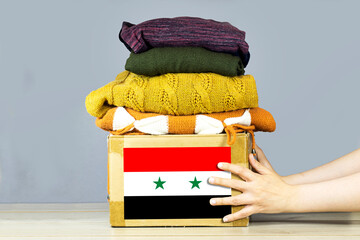 closeup woman hands holding carton box with used clothes for donation and Syria flag Earthquake, catastrophe volunteering